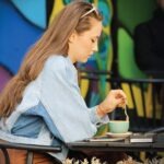 Woman sidewalk cafe stirring a cup of coffee, with an abstract multi-colored mural in the background.